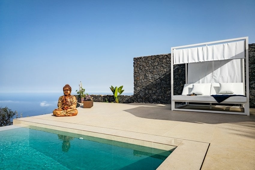 A serene outdoor space features a private infinity pool with clear water, bordered by a sun terrace. A comfortable lounge bed and a decorative buddha figure are positioned nearby, surrounded by plants and stone walls, creating a calming atmosphere with ocean views in the background.