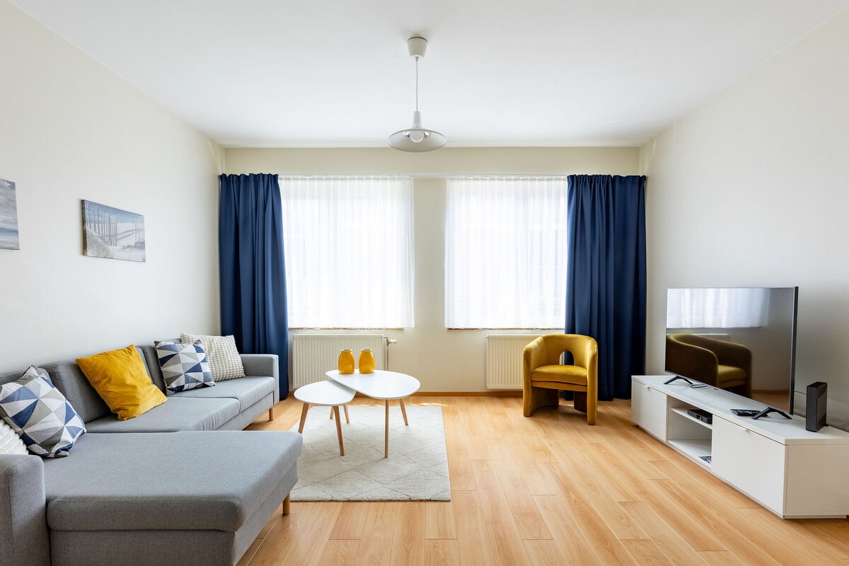 The living area features a light-colored sofa adorned with decorative pillows, complemented by a stylish armchair. A pair of round coffee tables sits in the center, surrounded by a soft area rug. Natural light enters through large windows dressed with sheer curtains and dark drapes.