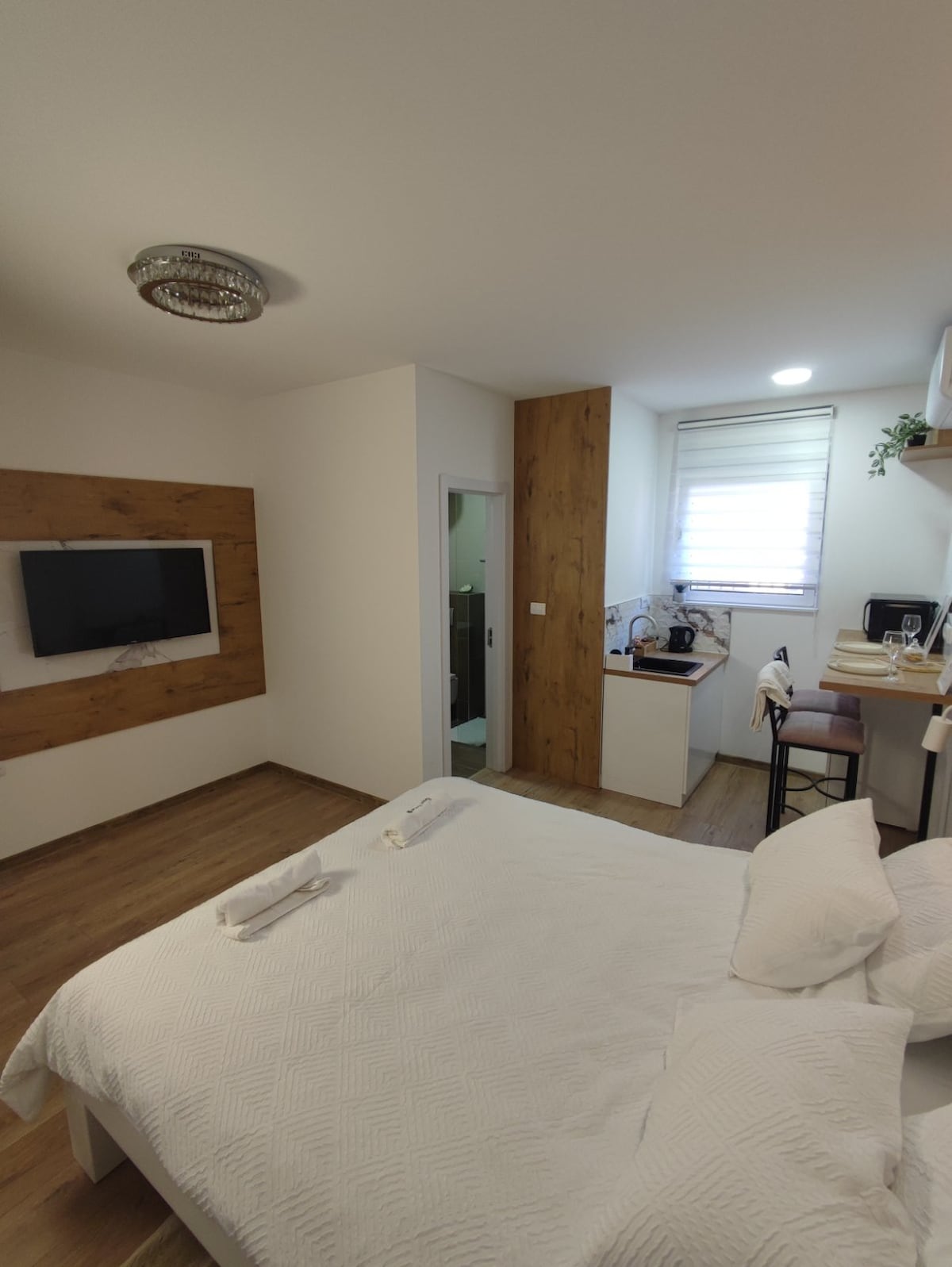 A modern accommodation space features a cozy sleeping area with a crisp white blanket atop a spacious bed. A wall-mounted TV is visible across from the bed. A kitchenette with essential appliances is situated in the corner, complemented by a small dining area with two chairs.