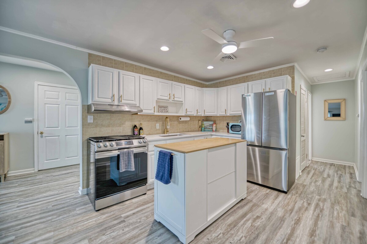 The kitchen features white cabinetry with a central island that includes a wooden countertop. Stainless steel appliances are positioned against a light-colored backsplash. A ceiling fan helps maintain airflow while natural light enters through the nearby entrance, enhancing the spaciousness of the area.