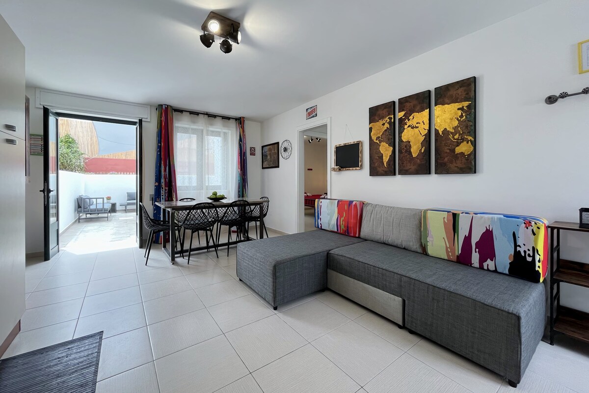 An open living area features a spacious gray sectional sofa, a dining table set for six, and colorful artwork depicting a world map. Natural light fills the room through large glass doors leading to an outdoor space, enhancing the inviting atmosphere.