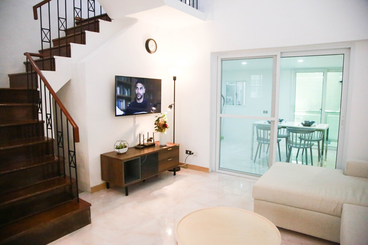 The living area displays a contemporary design with a comfortable sectional sofa and a wooden TV cabinet. A flat-screen television is mounted on the wall. Natural light fills the space through large windows, while a staircase leads to the upper level.