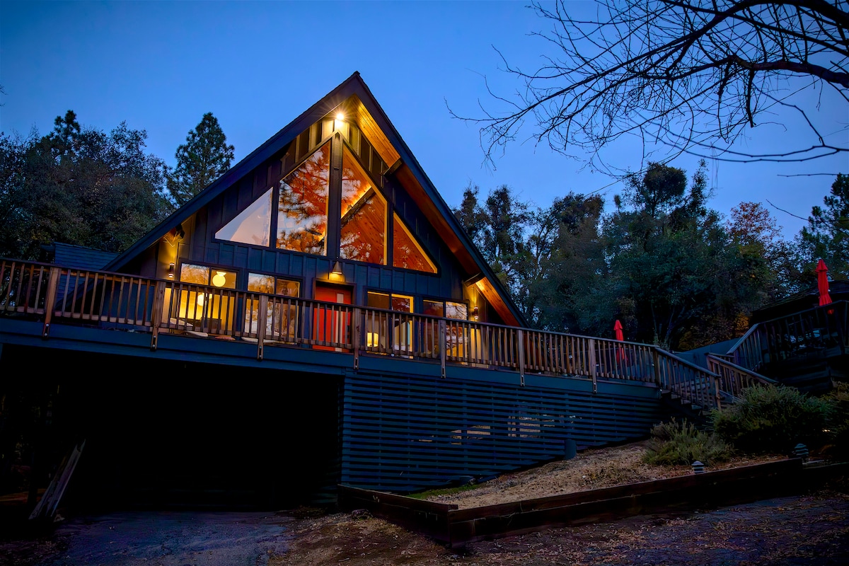 The A-Frame cabin is viewed from the front at twilight, highlighting its distinctive triangular design and large windows. The illuminated interior offers a glimpse of the cozy ambiance inside. The surrounding nature and deck are visible, creating a serene outdoor setting.