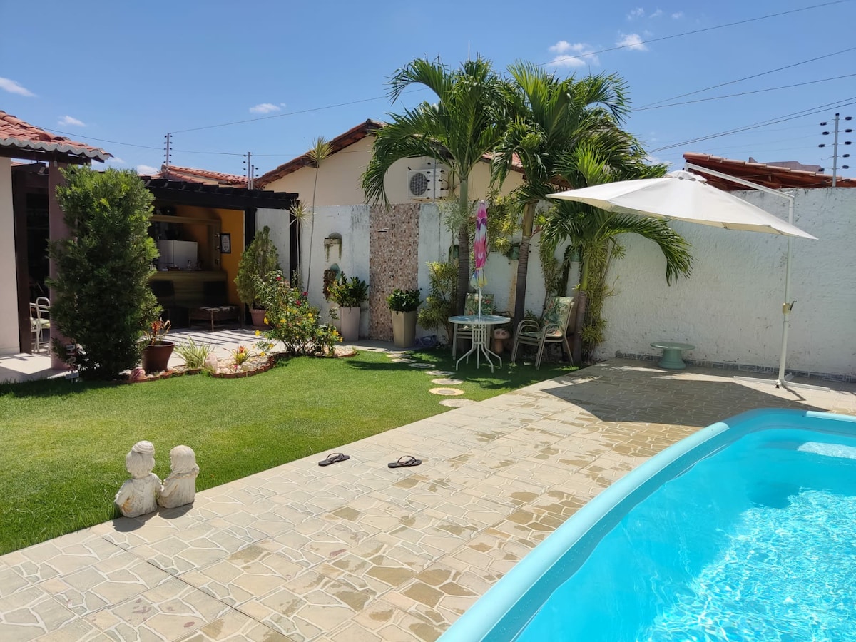 A private outdoor space is featured, showcasing a clear swimming pool surrounded by well-maintained grass. Tropical plants are located near the pool, and a shaded seating area with a table and chairs is visible, offering a space for relaxation.