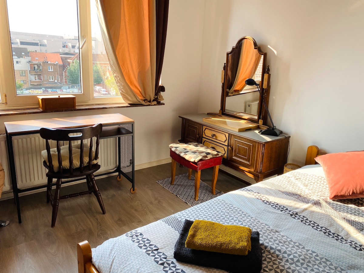 The image showcases a well-lit bedroom featuring a spacious bed with patterned bedding. A wooden desk and chair are positioned beside a vintage dresser, complemented by a small upholstered stool. Natural light filters through the window, illuminating the warm tones of the room.