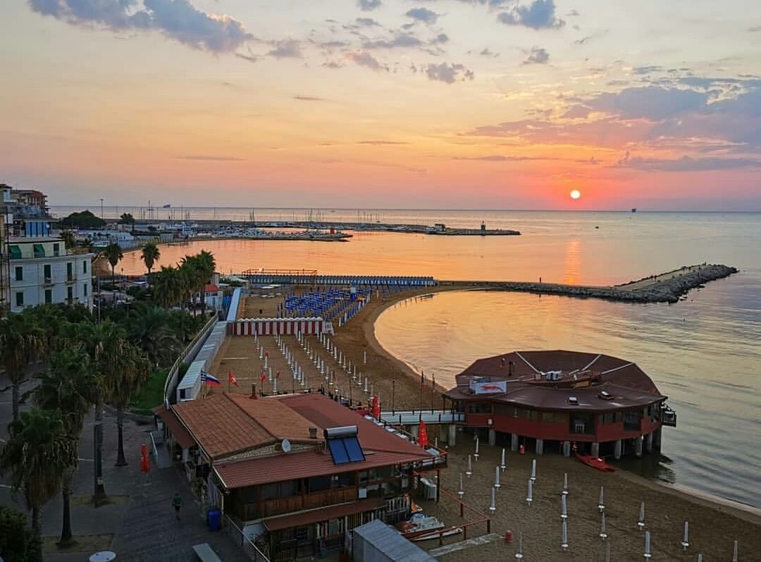 A beach scene is captured at sunset, with calm waters reflecting warm hues. A sandy shoreline curves gently, lined with umbrellas and lounge chairs. A beach establishment features a terracotta roof, while palm trees dot the area, contributing to a relaxed coastal atmosphere.