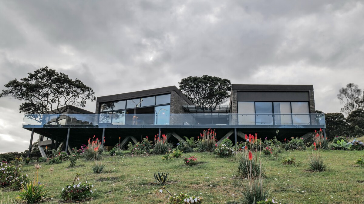 A modern house is positioned on a hillside, featuring large glass windows that provide panoramic views. The structure is supported by beams and is surrounded by a landscaped garden with flowering plants and greenery, creating a harmonious connection with nature.