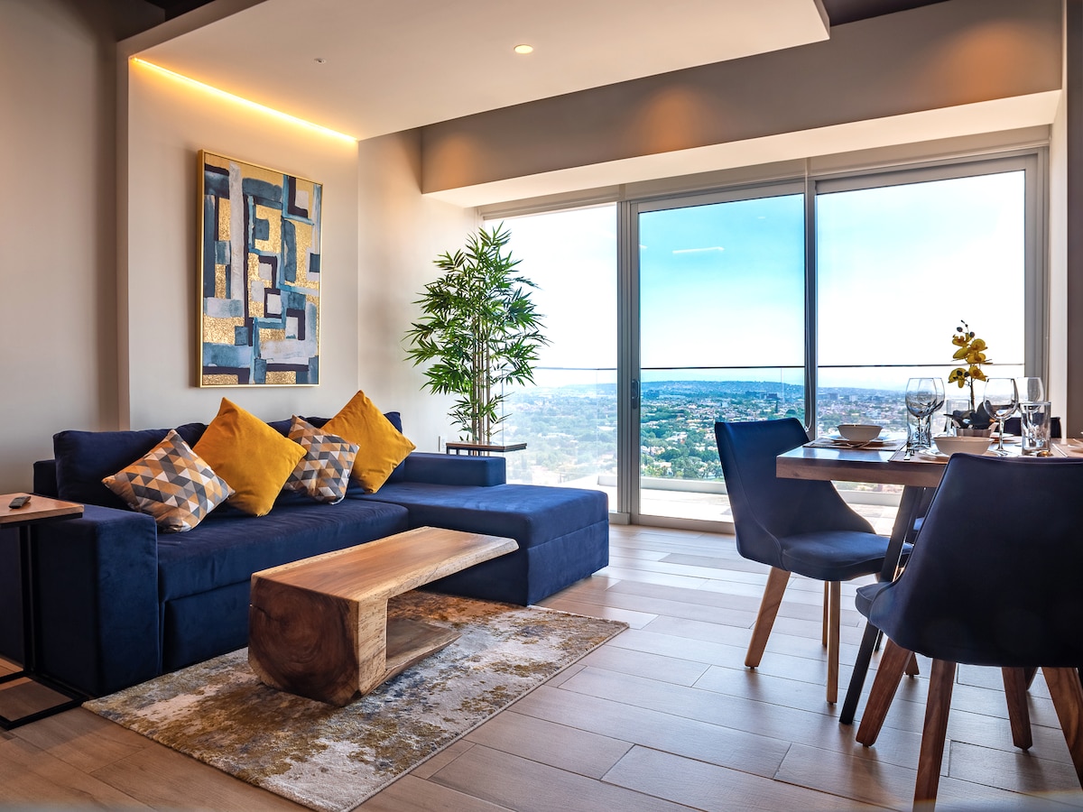 An inviting living area features a blue sofa with decorative pillows and a wooden coffee table. A dining table with elegant table settings is positioned nearby. Large windows allow for ample natural light, offering a sweeping view of the city skyline.