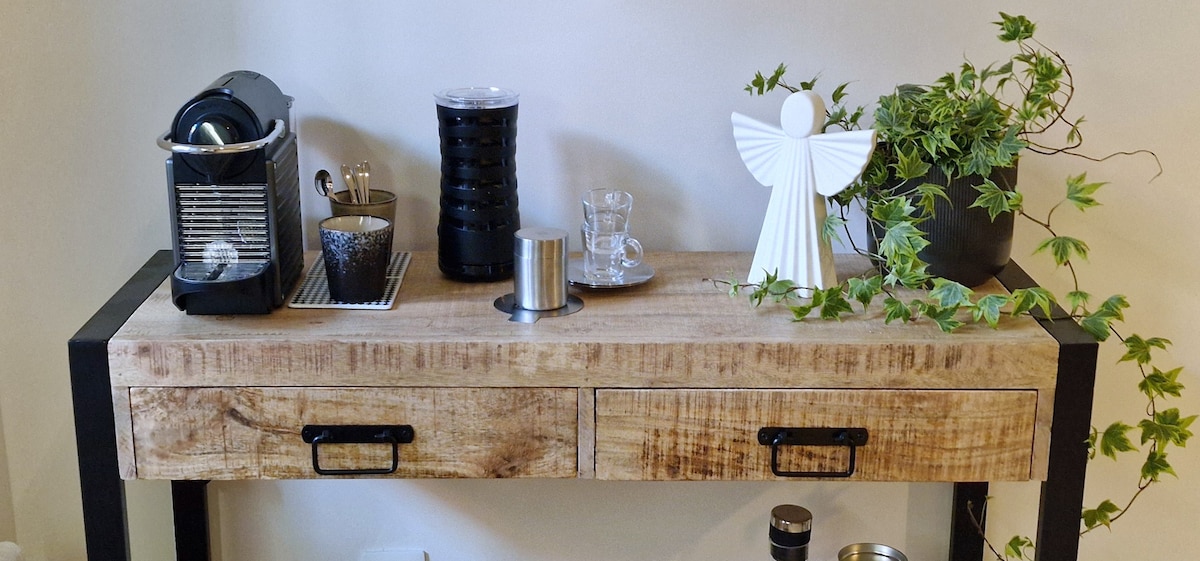 A coffee corner is presented with a modern wooden console featuring two drawers. A Nespresso machine, a French press, and glassware are neatly arranged on top, along with a decorative angel figurine and a potted plant, enhancing the inviting space.