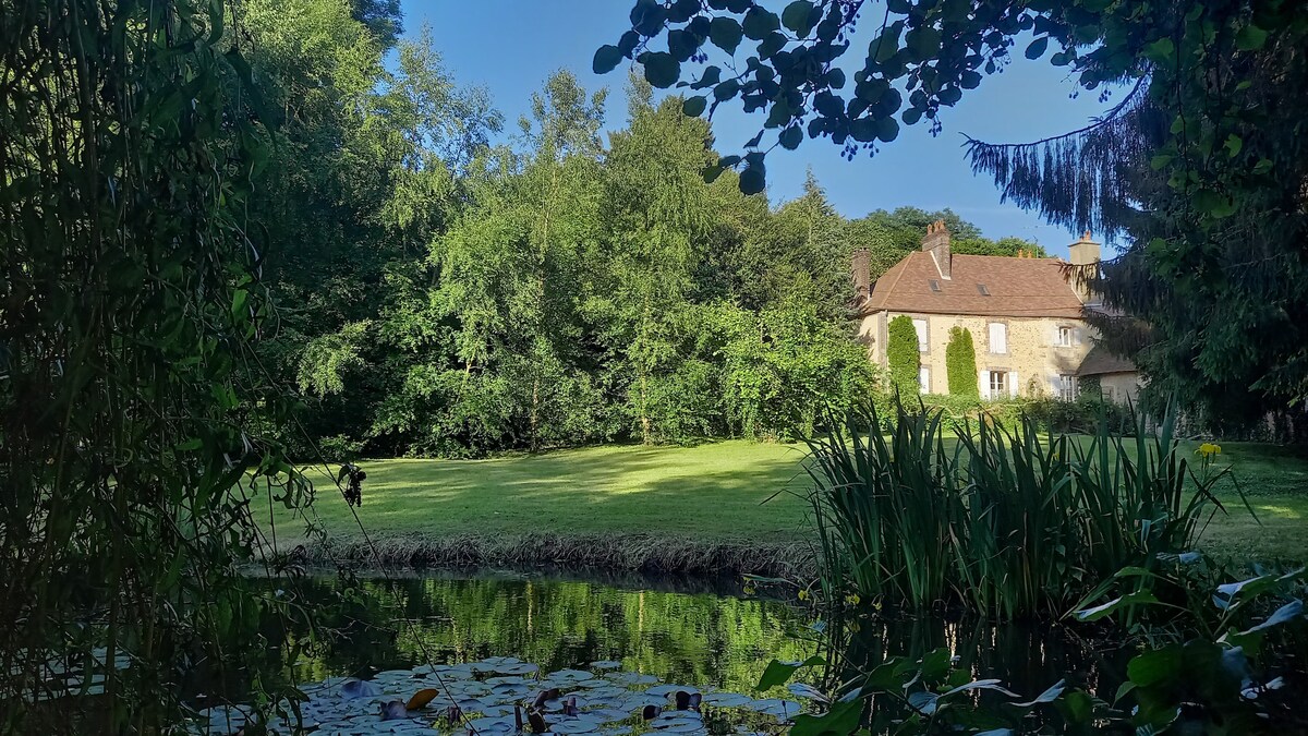 A serene scene captures a lush green landscape framed by foliage. A well-maintained garden is visible alongside a reflective pond, with a charming house partially obscured by trees in the background, showcasing a blend of natural and architectural beauty.