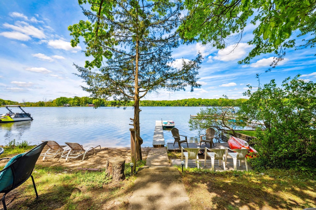 The serene shoreline of Pickerel Lake is featured, showcasing a sandy beach area with several chairs positioned for relaxation. A private dock extends into the water, accompanied by two kayaks and a vibrant paddleboat. Lush green trees frame the calming scene under a clear blue sky.