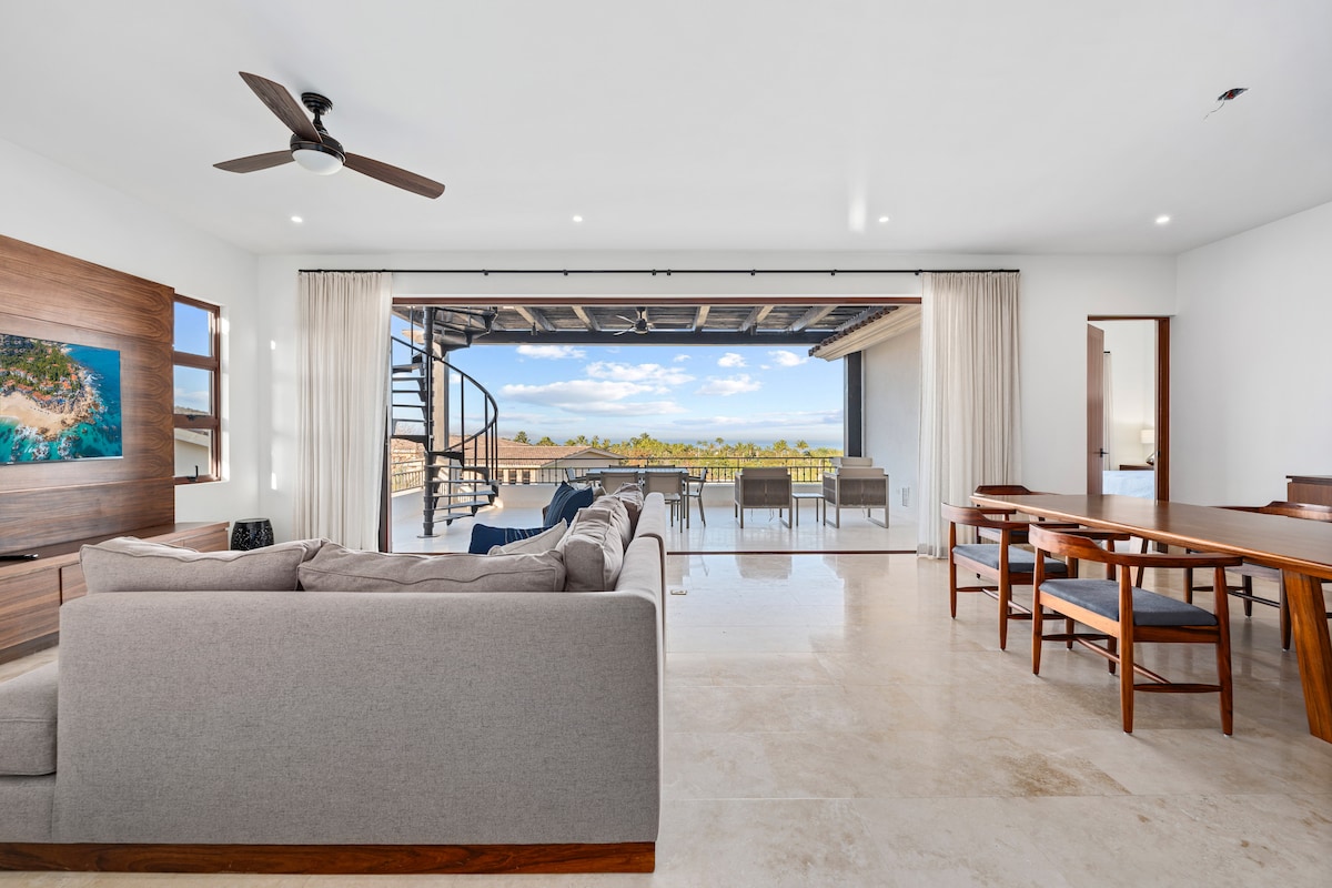 A spacious living area is presented, featuring a comfortable sectional sofa and a wooden dining table. Large glass sliding doors offer a view of the outdoor dining space and ocean beyond. Natural light brightens the room, while a decorative wall adds warmth to the modern aesthetic.