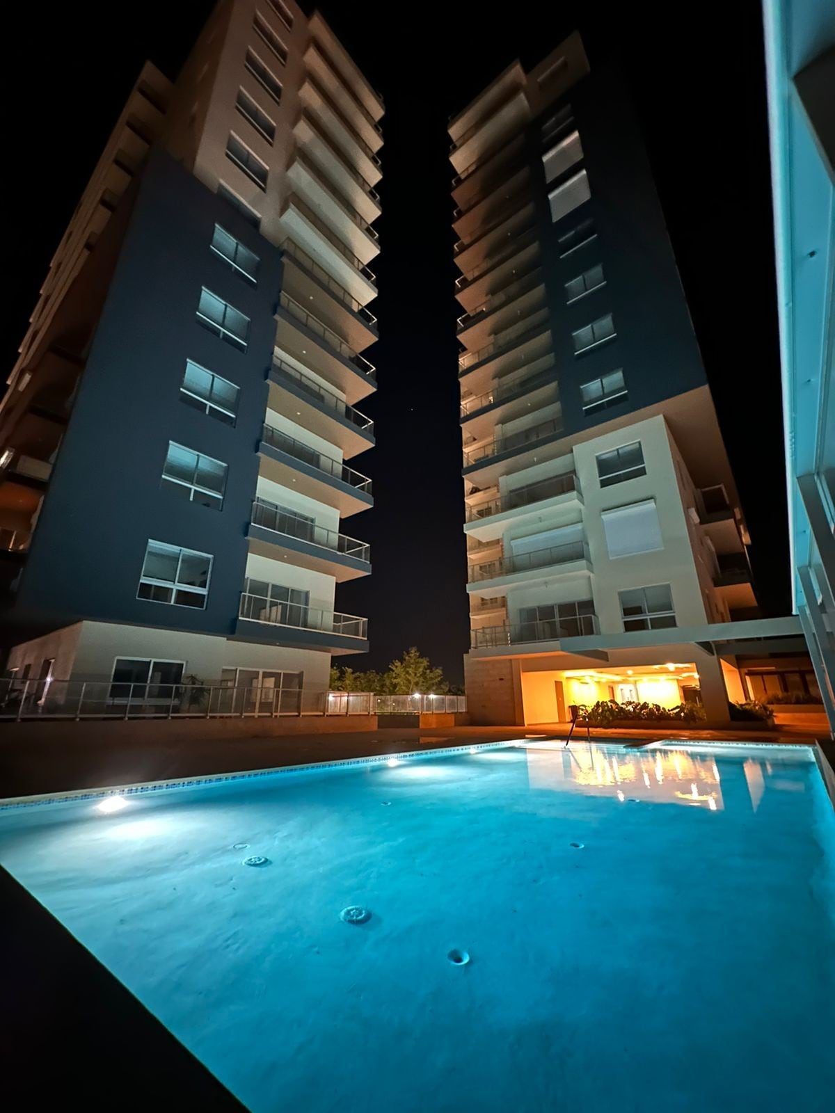 Two tall buildings are depicted at night with bright lights illuminating their façades. A resort-style pool is visible in the foreground, reflecting the surrounding lights. The tranquil water enhances the serene atmosphere of the setting.