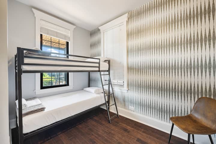 غرفة نوم بسرير معدني بطابقين وورق حائط منقوش وكرسي خشبي ونافذة مع ستائر.