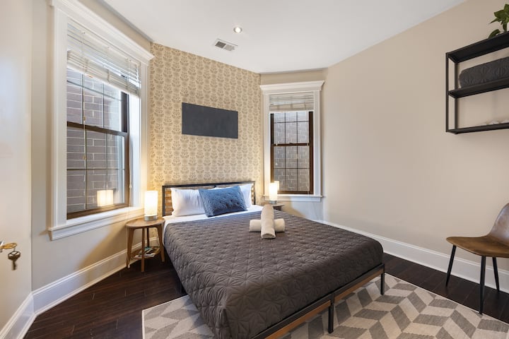 غرفة نوم بسرير متوسط الحجم، وورق حائط منقوش، ونافذتان مع ستائر، وسجادة هندسية على الأرض.