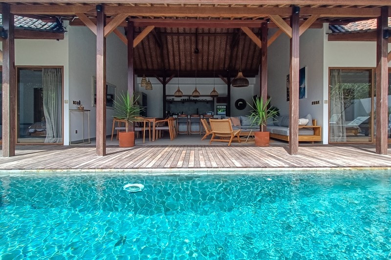 A refreshing swimming pool is visible in the foreground, with clear water reflecting the surrounding wooden structure. The open living space features a kitchen, dining area, and relaxation zone, all enhanced by natural light streaming through expansive glass doors.