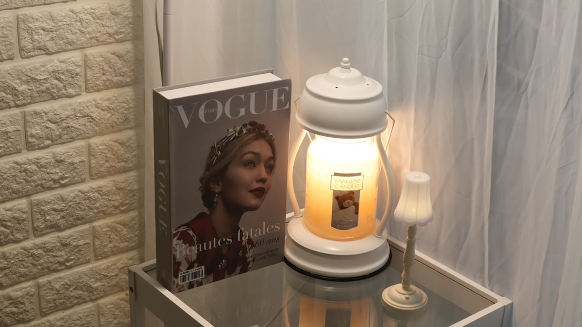 A bedside table is featured, holding a decorative lamp and a small candle. A fashion magazine lies next to the lamp, with soft lighting creating a warm ambiance. The backdrop consists of textured wall panels and sheer white curtains.