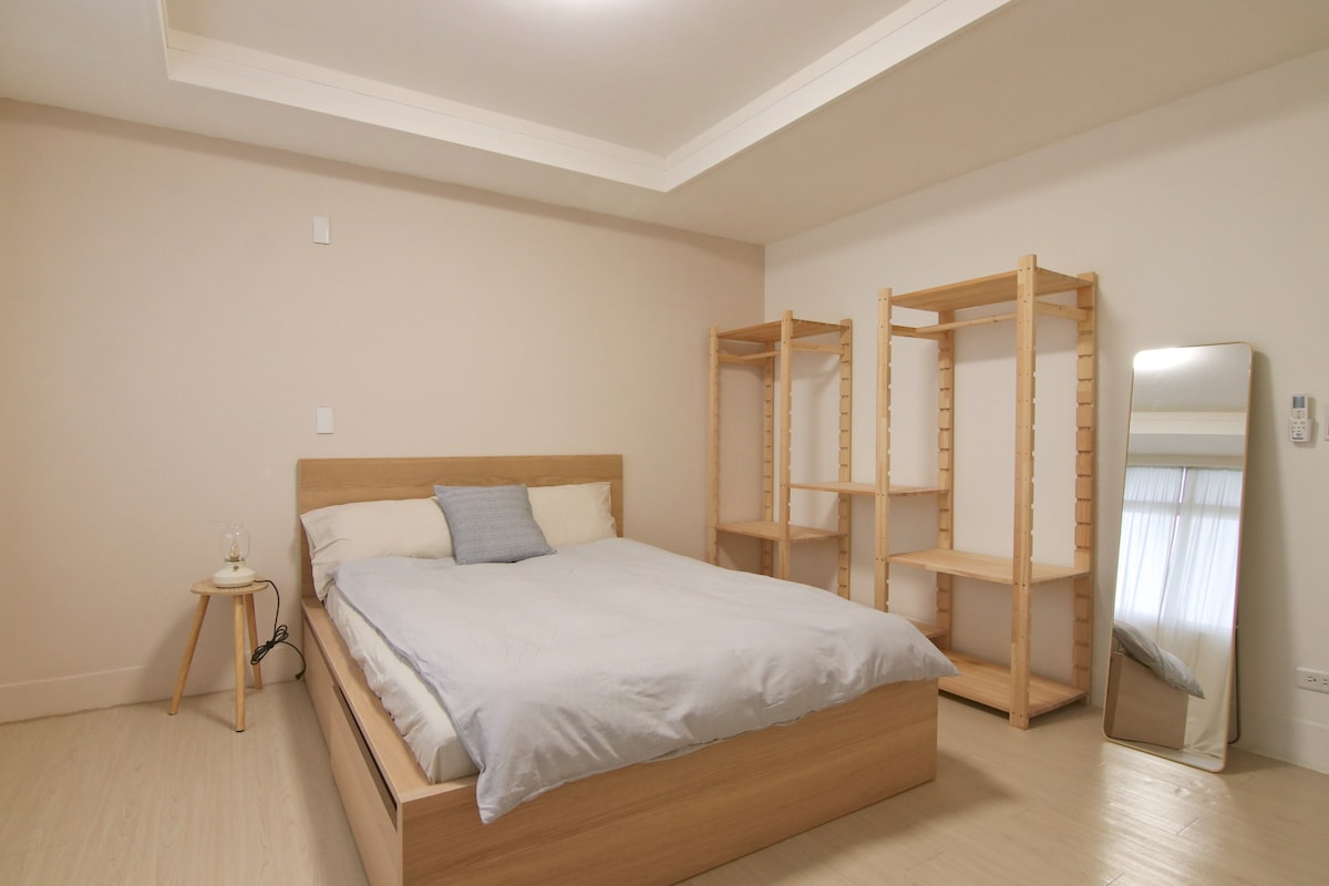 A spacious room features a wooden bed with a light duvet, complemented by a single accent pillow. A mirror stands against the wall, and open shelving provides storage options. Soft natural light enters through a window, enhancing the calming atmosphere.