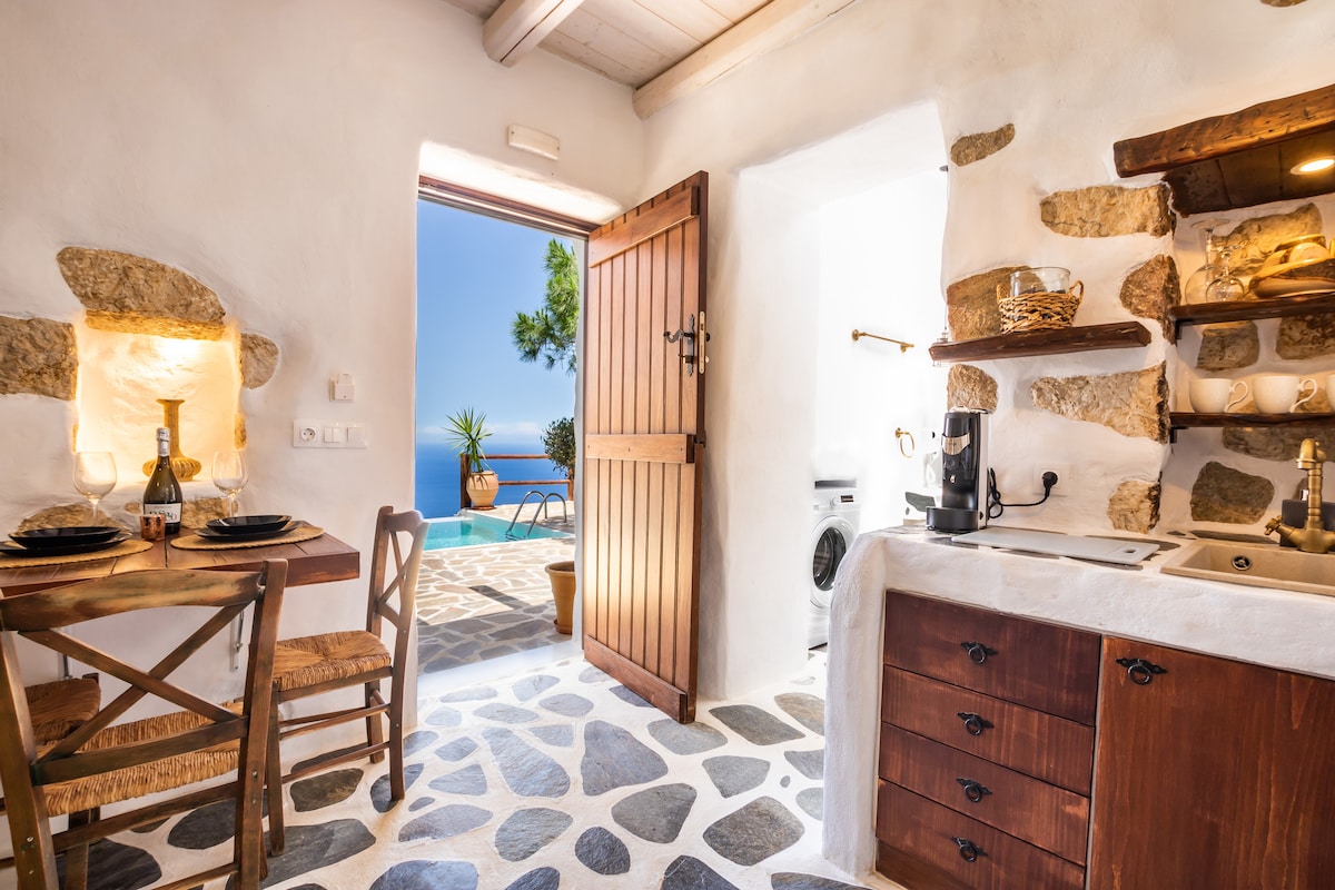 A well-equipped kitchen is shown with wooden cabinetry and a washing machine. A small dining table set for two is visible, with views of the private swimming pool and the Aegean Sea beyond. Natural light enters through an open door, highlighting the stonework.
