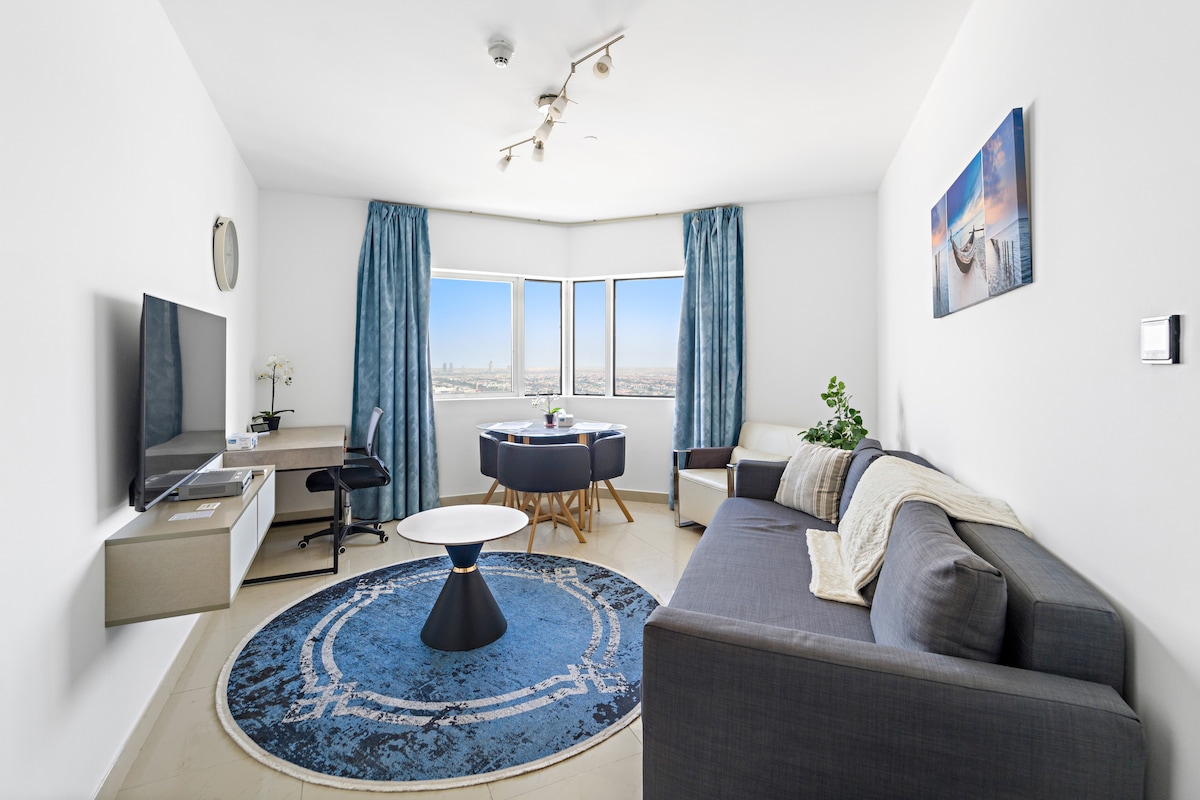 A modern living area features large windows offering sweeping views of the city skyline. A plush gray sofa and a circular coffee table sit on a decorative rug. A desk with a chair is positioned near the window, creating an inviting workspace. Natural light illuminates the room.