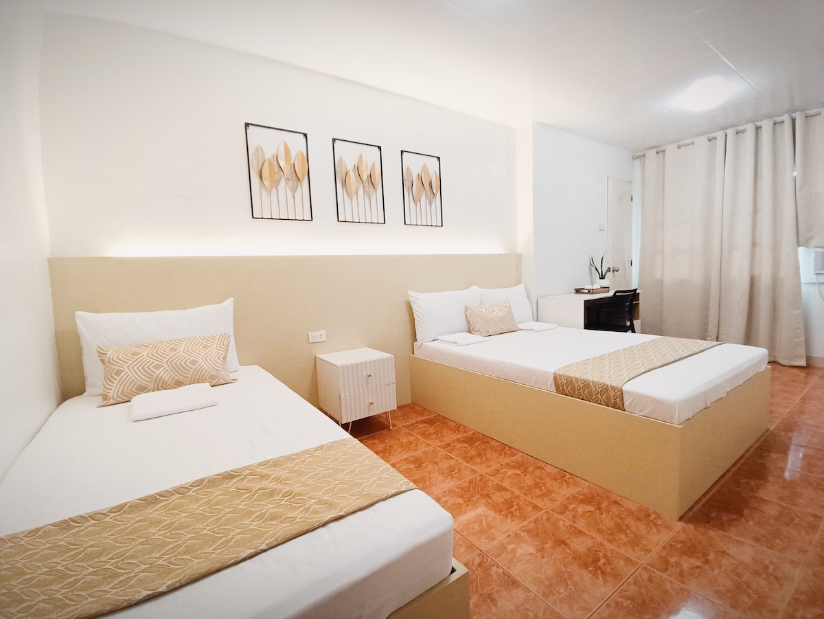 A guest room is presented with two beds, one double and one single. A soft beige and white color scheme is utilized, complemented by decorative wall art. A work desk is situated in the corner, while light filters in through sheer curtains.