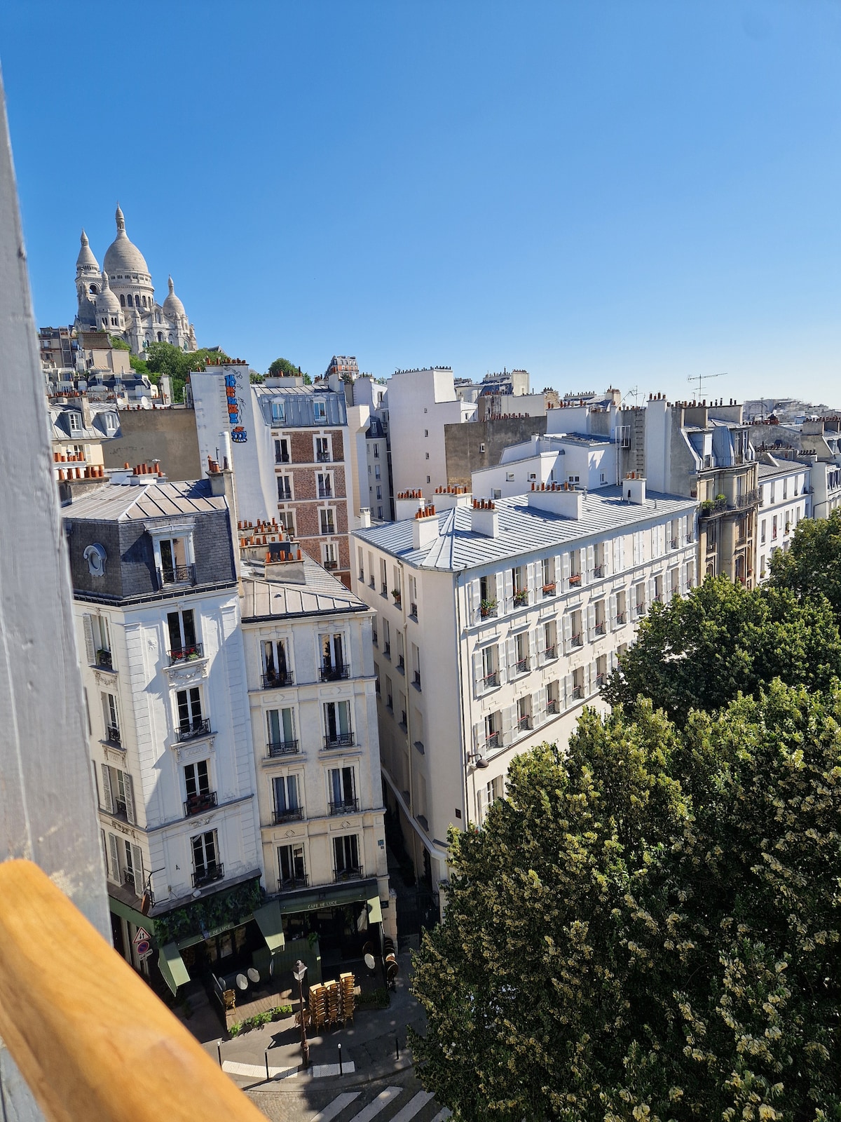 A view from the balcony reveals the charming rooftops of Montmartre and the distant Sacré-Cœur, framed by lush green trees. The scene captures the essence of the vibrant neighborhood, with a clear blue sky enhancing the inviting atmosphere of the surroundings.