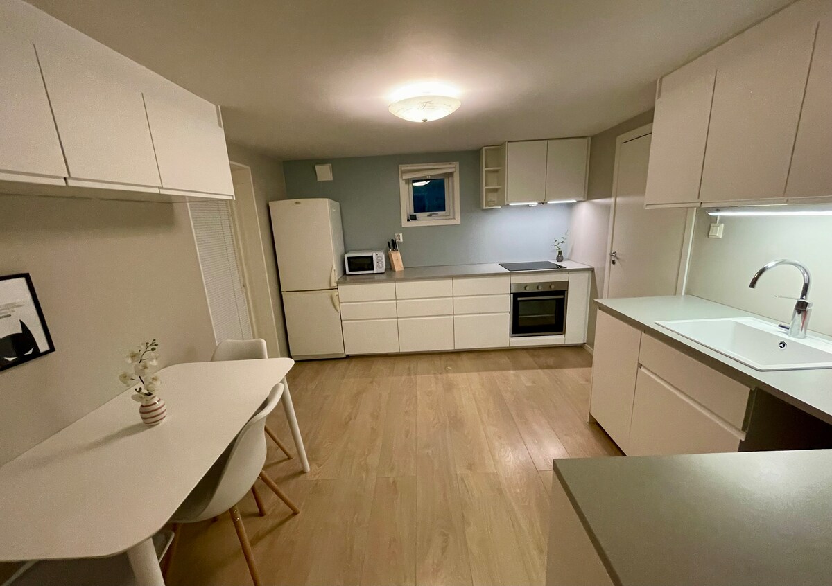 The kitchen is designed with modern white cabinetry, including overhead and base units. A dining table with light-colored chairs accommodates four. The kitchen is equipped with an oven, a refrigerator, and a microwave, complemented by well-placed lighting and a window that invites natural light.