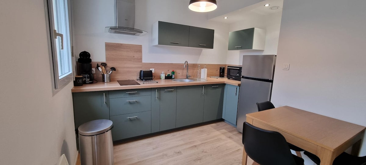 A modern kitchen is equipped with essential appliances, including a refrigerator and a microwave. The cabinetry features a combination of muted green and natural wood tones. A small dining table with two black chairs sits nearby, creating a functional and inviting cooking space.