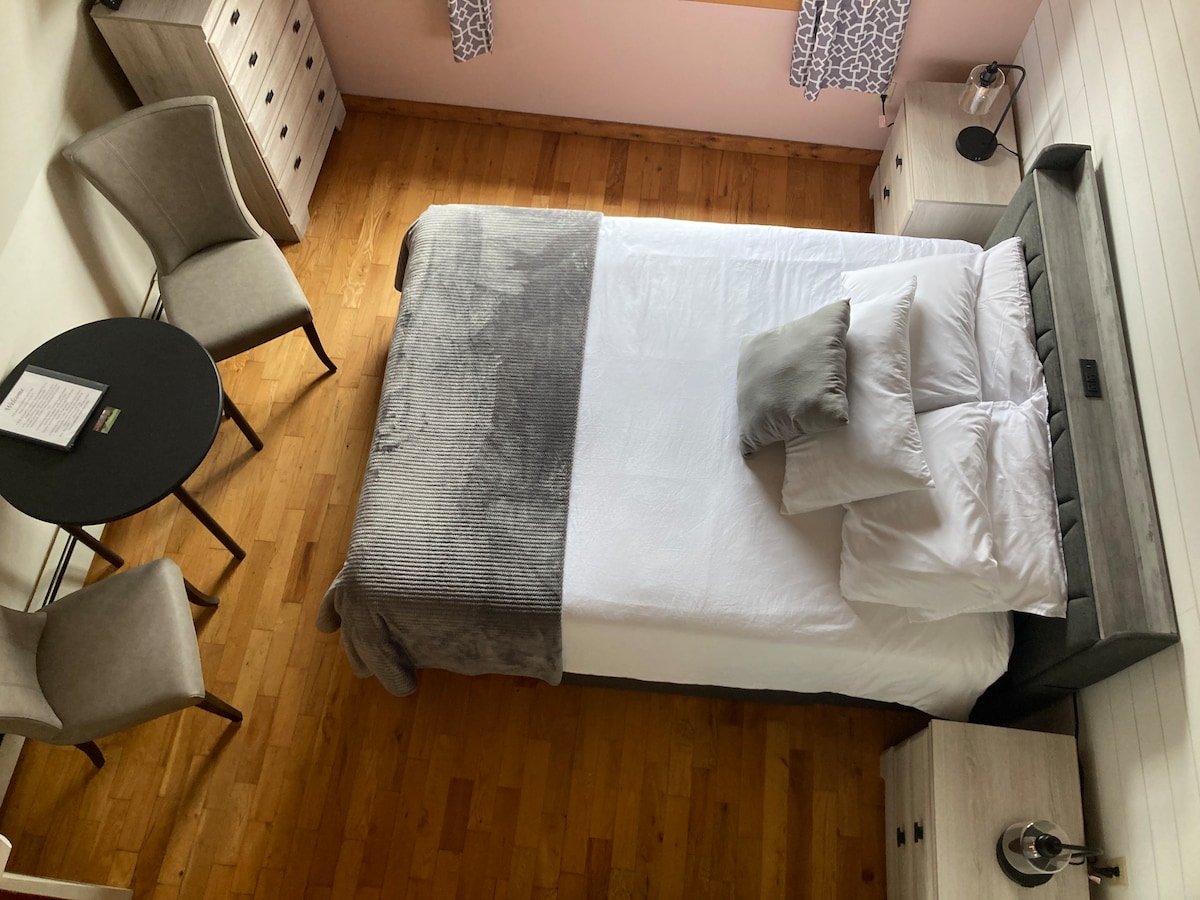 A welcoming sleeping area features a spacious bed with soft bedding and decorative pillows. Two modern bedside tables with lamps are placed on either side. A small round table and two chairs are located nearby, all set against warm wooden flooring.