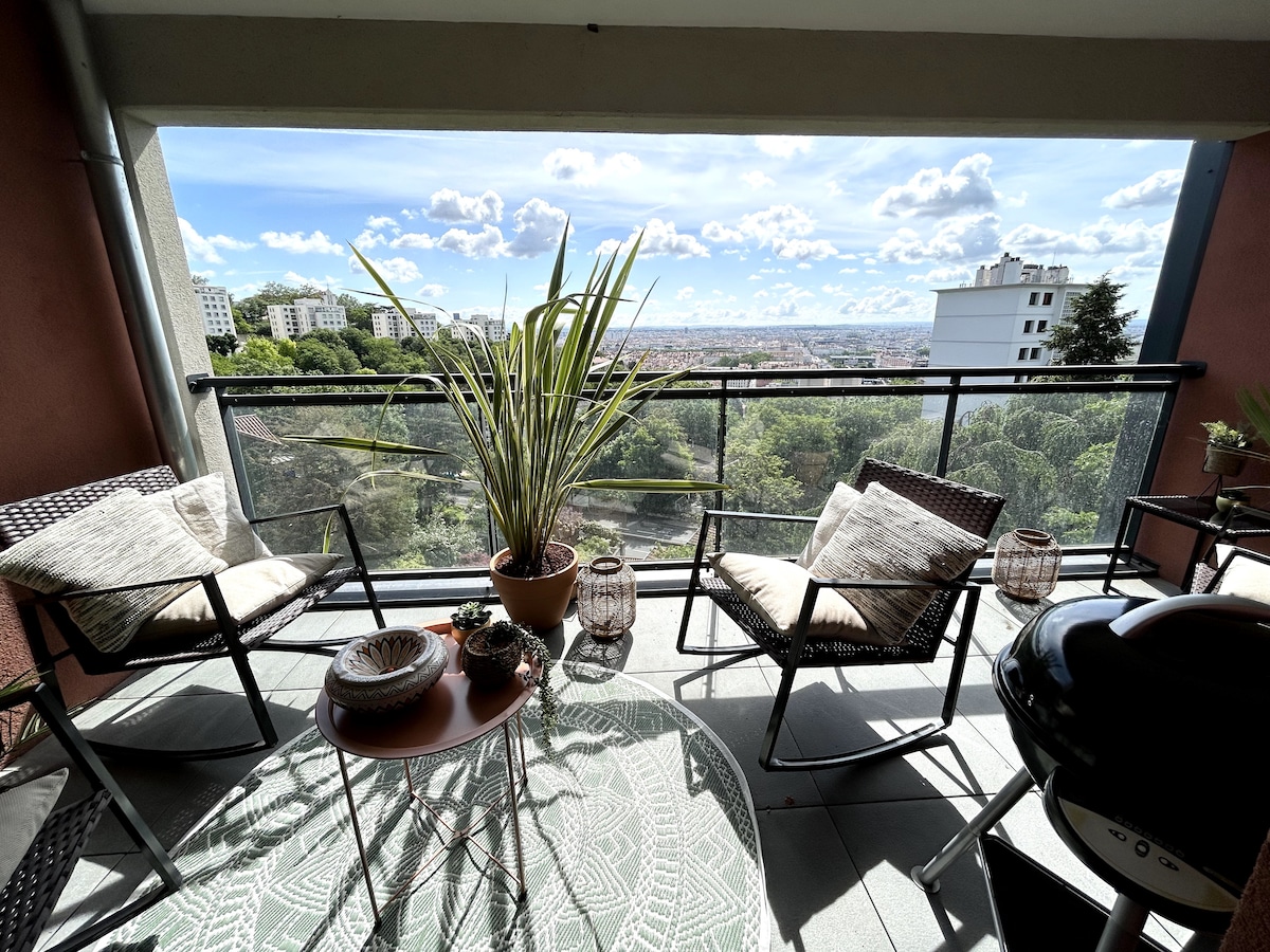 An inviting balcony features two woven chairs and a small wooden table, complemented by decorative plants. A barbecue grill is positioned nearby, and a patterned outdoor rug adds texture to the floor. The view extends across the green landscape and city skyline.