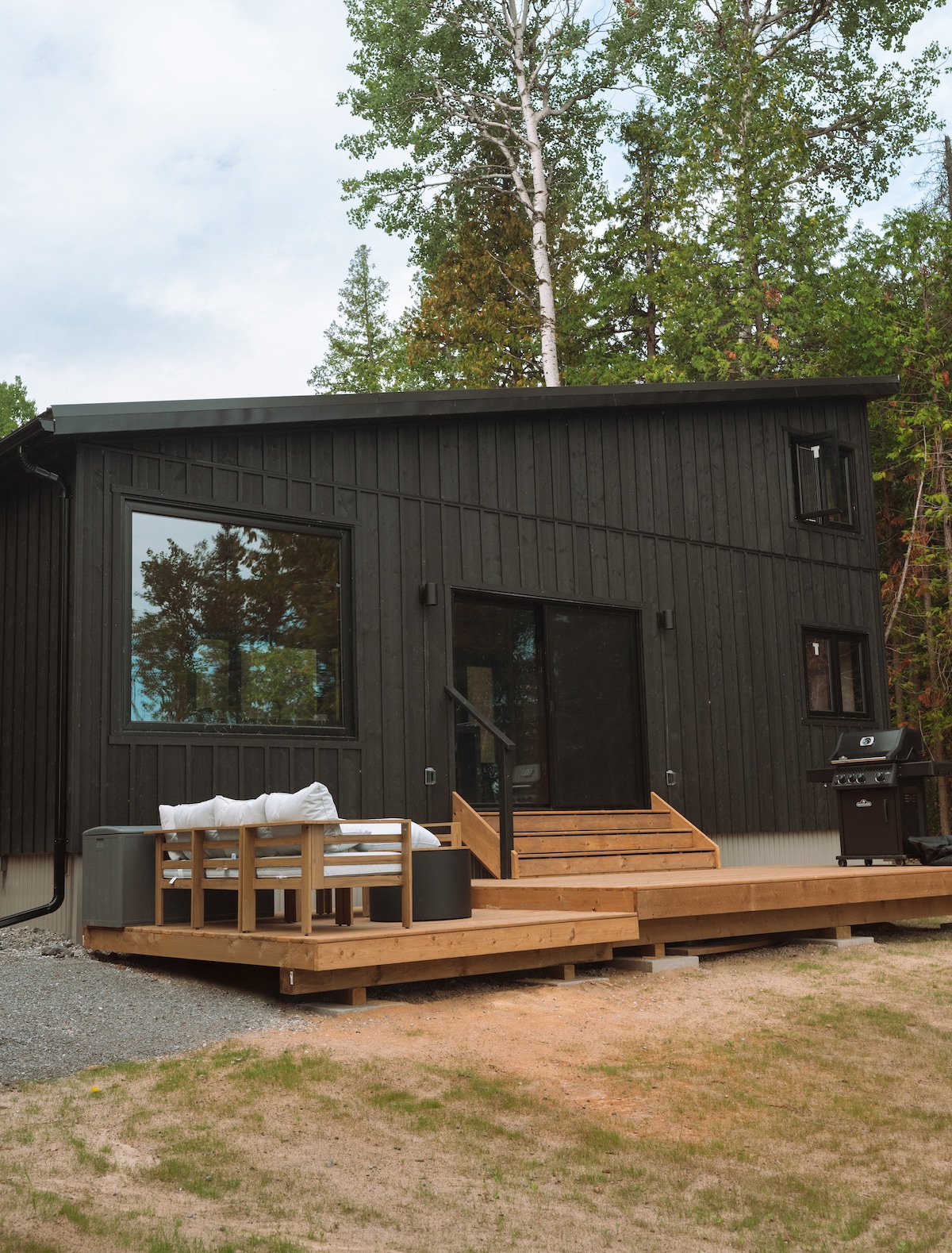 A modern cabin exterior is featured, with dark wood siding and large windows. A wooden deck with steps leads to a comfortable outdoor seating area, alongside a BBQ grill, all surrounded by lush greenery.