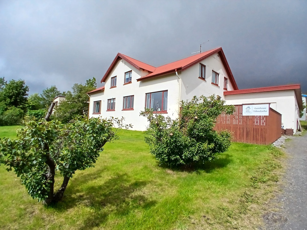 A charming guesthouse is presented with a red-roofed exterior and multiple windows. The surrounding green lawn features small trees, enhancing the inviting appearance. A sign is visible, indicating the guesthouse's name, while a partially cloudy sky provides a contrasting backdrop.
