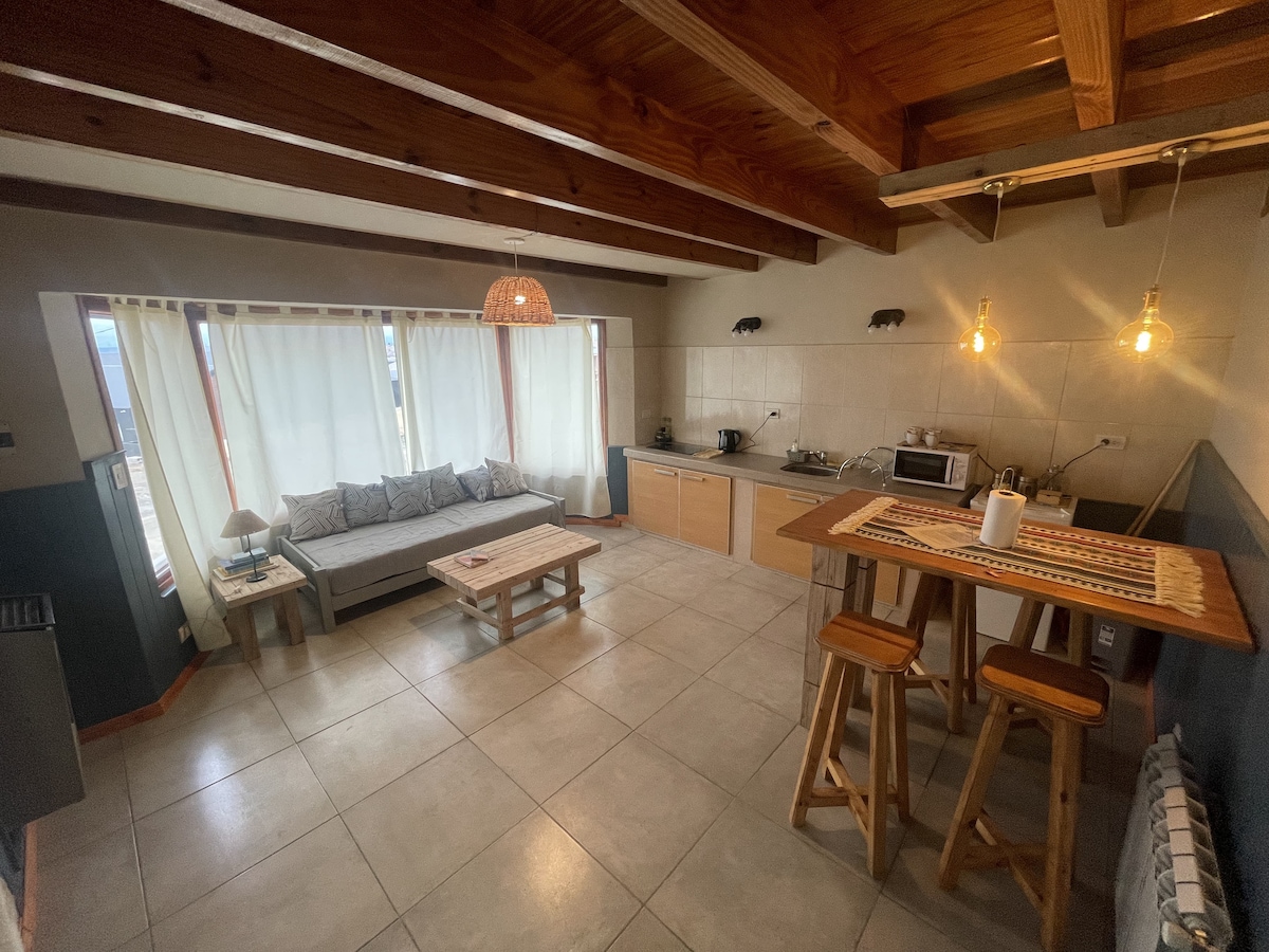 A modern kitchen and living area display open-plan layout features. A comfortable sofa sits alongside a rustic wooden table, with natural light filtering through sheer curtains. Warm wooden beams draw the eye upward, while practical kitchen appliances are neatly arranged for convenience.