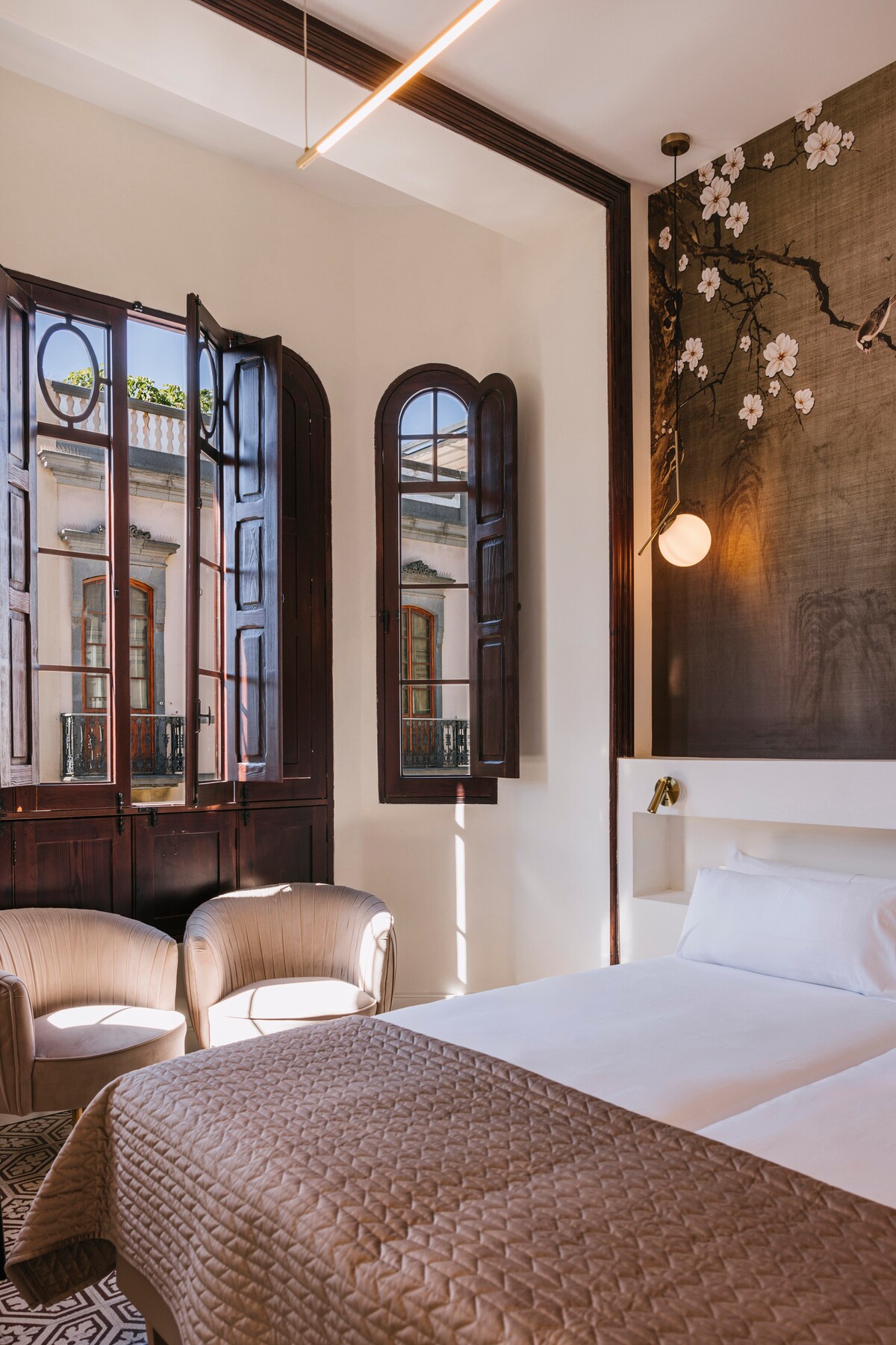 A well-lit room features large, open windows adorned with wooden shutters, allowing natural light to fill the space. Two circular chairs are positioned beside a spacious bed, which is complemented by a decorative wall featuring floral artwork.