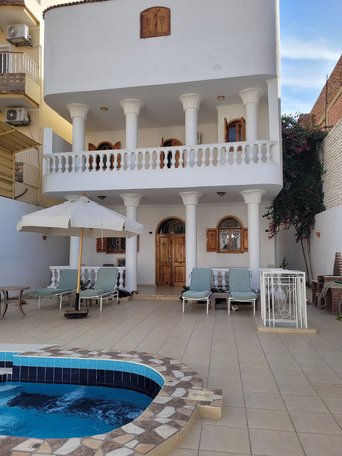 A two-story building is featured with arched doorways and elegant columns. A patio contains lounge chairs and an umbrella, while a small pool, with a mosaic border, adds a refreshing touch to the outdoor space.