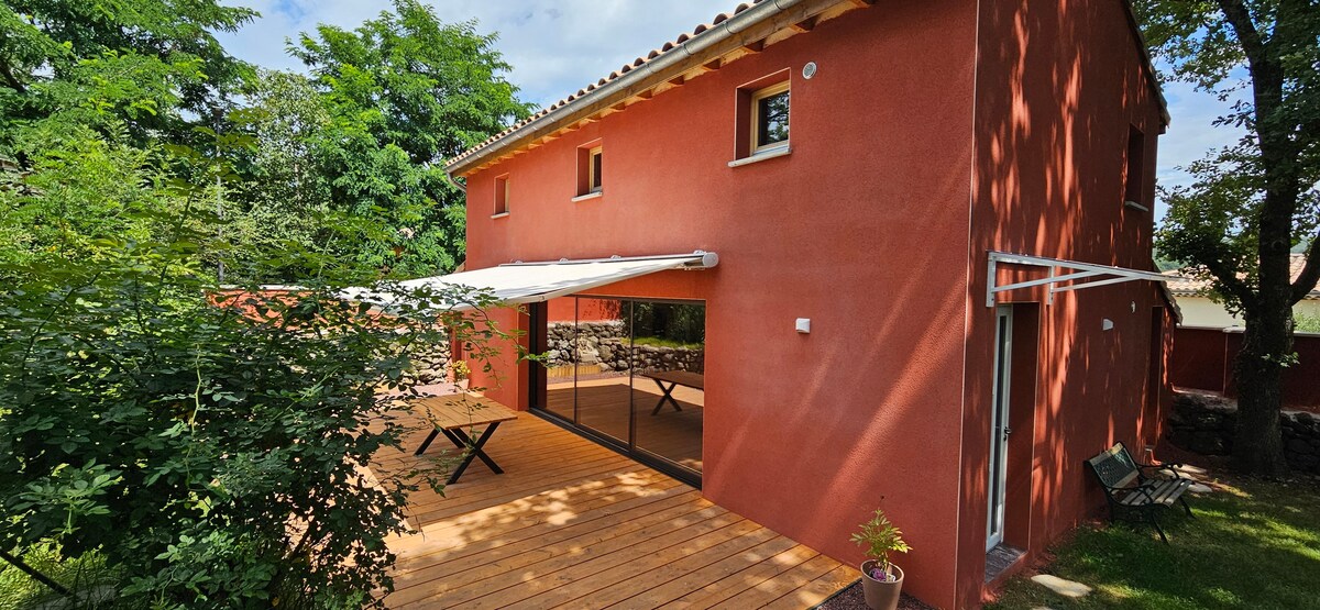 A modern house exterior is highlighted, showcasing a warm red facade and large sliding glass doors. A spacious wooden terrace extends from the house, featuring a shaded outdoor dining area. Surrounding foliage provides a sense of privacy and natural beauty.