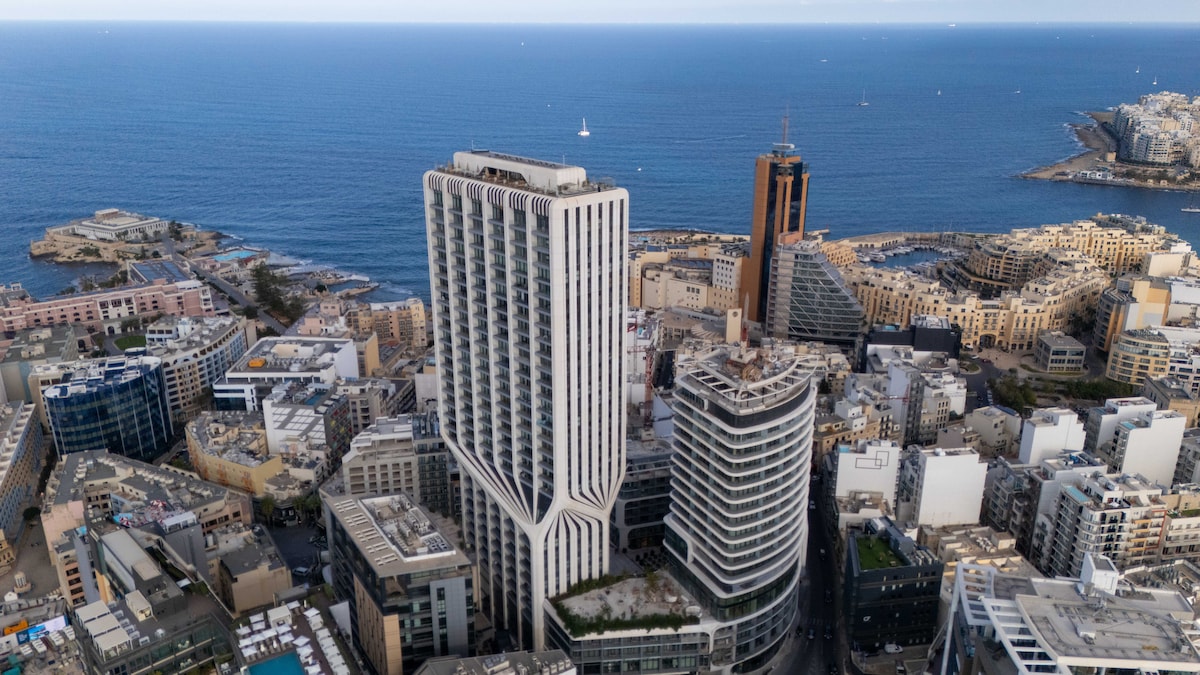 Aerial view showcases the contemporary architecture of Mercury Towers, rising prominently near the coastline. The surrounding urban landscape is visible, with a blend of modern buildings and the shimmering blue sea in the background, creating an inviting atmosphere for visitors.
