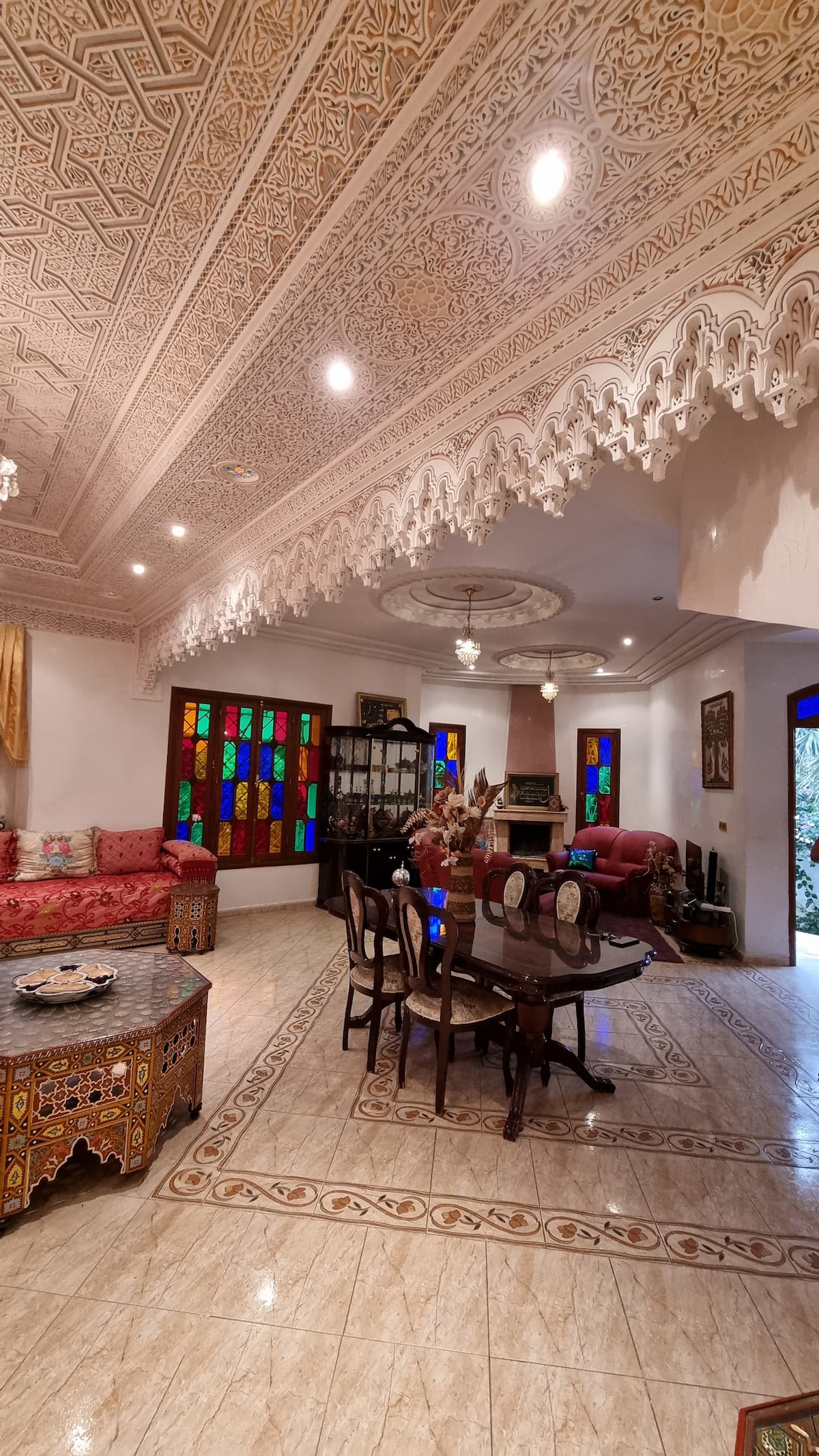 The interior space features intricately carved ceilings and colorful stained glass windows, adding character to the area. A dining table is surrounded by wooden chairs, while various seating arrangements are placed against the walls. Natural light enhances the elegant tile flooring.