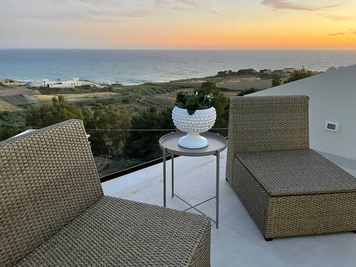 An outdoor terrace features two woven lounge chairs and a small table. A decorative white pot rests on the table, holding greenery. The open sky transitions into warm sunset hues above a distant view of the ocean and coastline.