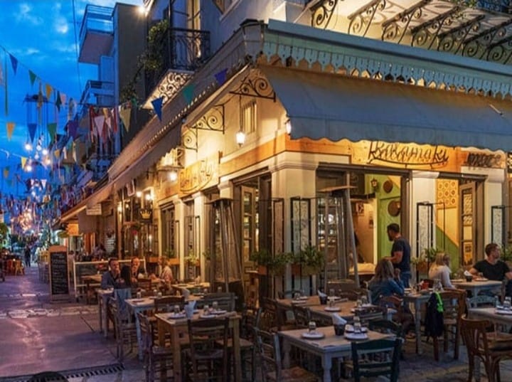 An outdoor dining area is set along a lively street, featuring tables adorned with white tableware. Vibrant decorations and colorful bunting add a festive touch. Patrons engage in conversation while enjoying their meals, with soft lighting creating a welcoming ambiance as evening approaches.
