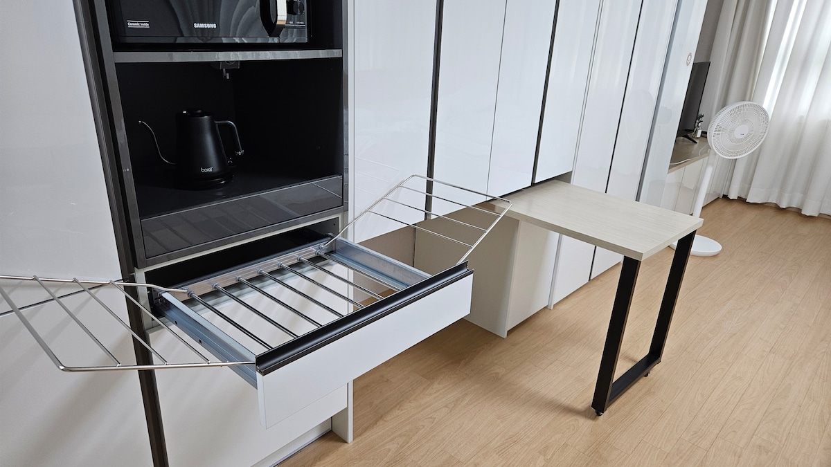 A modern kitchen area is visible, featuring a sleek refrigerator with an open drawer showcasing a pull-out oven rack. A small table is positioned nearby, complementing the minimalist design. Natural light filters through sheer curtains, creating a bright and open ambiance.