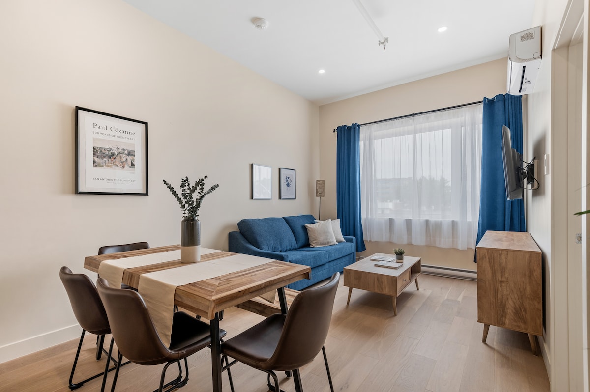 A bright living area features a blue sofa and a coffee table set on light hardwood flooring. A dining table with four chairs is positioned nearby. Natural light is filtered through a large window with blue curtains, enhancing the minimalist decor.