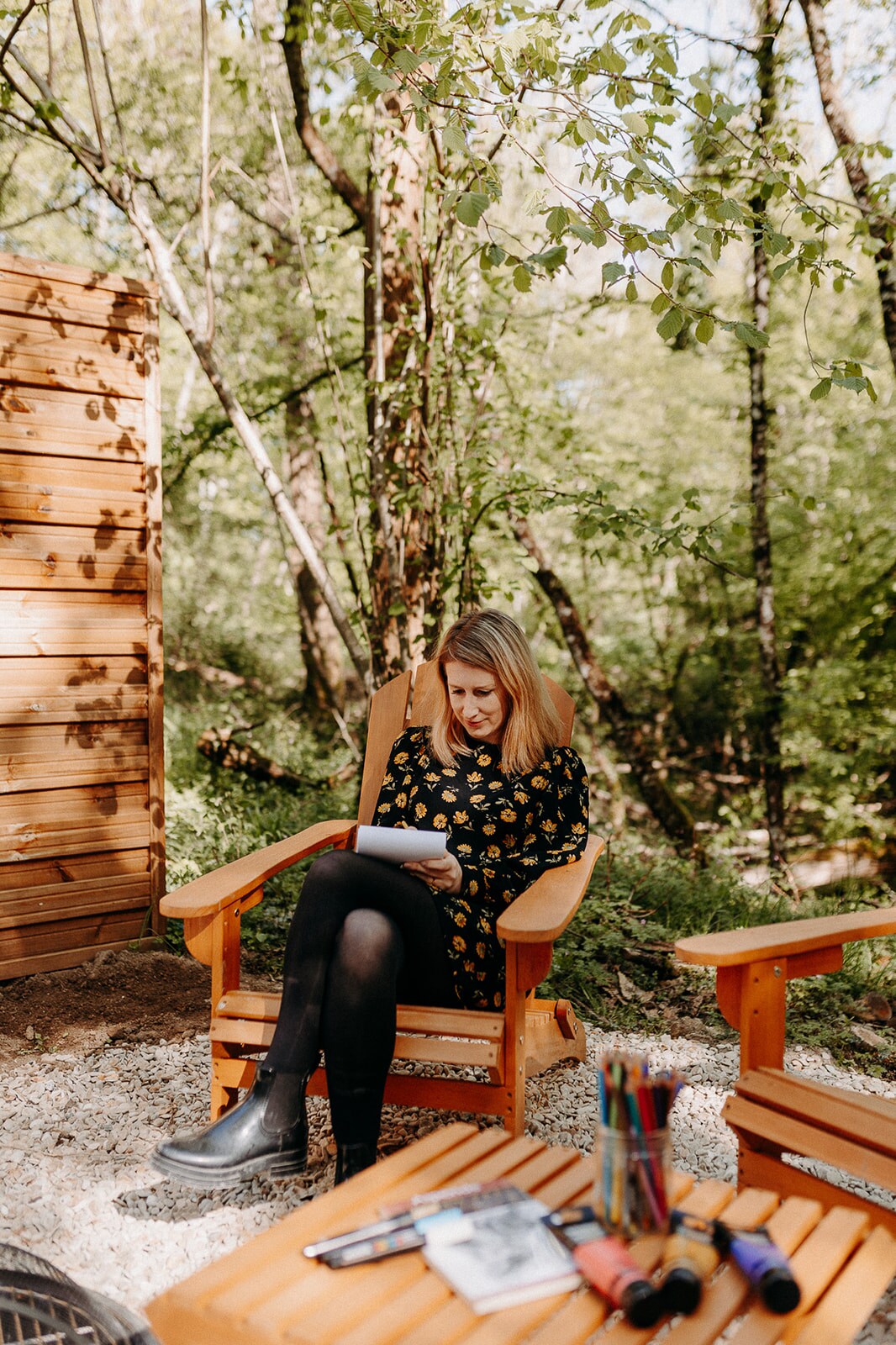 A serene outdoor space features two wooden Adirondack chairs amidst a lush forest setting. A small table hosts various colorful pens and markers. Sunlight filters through the leaves, casting gentle shadows on the gravel surface, creating a peaceful area for relaxation or creative pursuits.