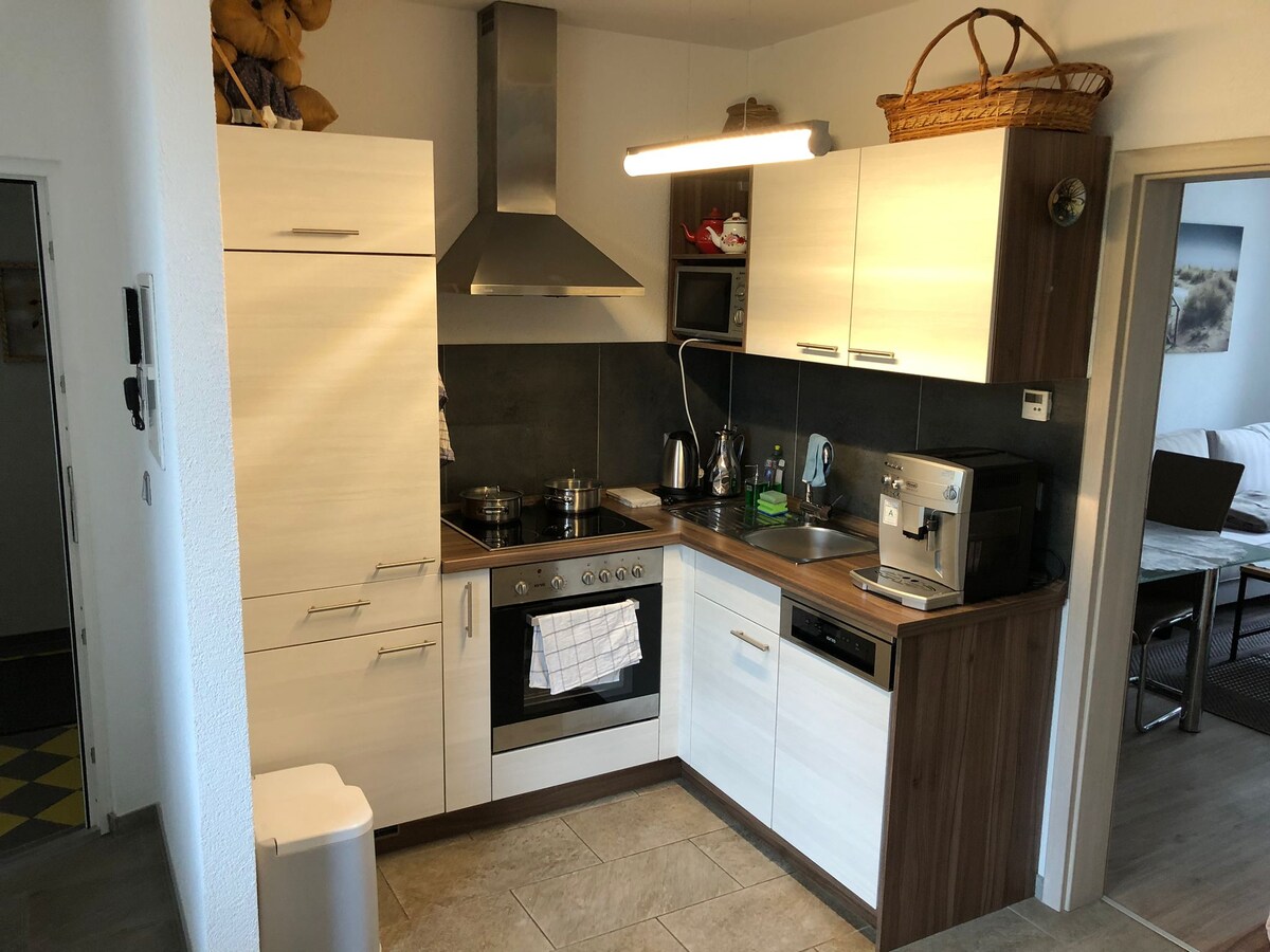 A compact kitchen area features modern cabinetry in light tones and wooden accents. Stainless steel appliances, including a stove and an espresso machine, are prominently displayed. A small microwave and kettle complement the workspace, with a light fixture providing illumination above the countertop.