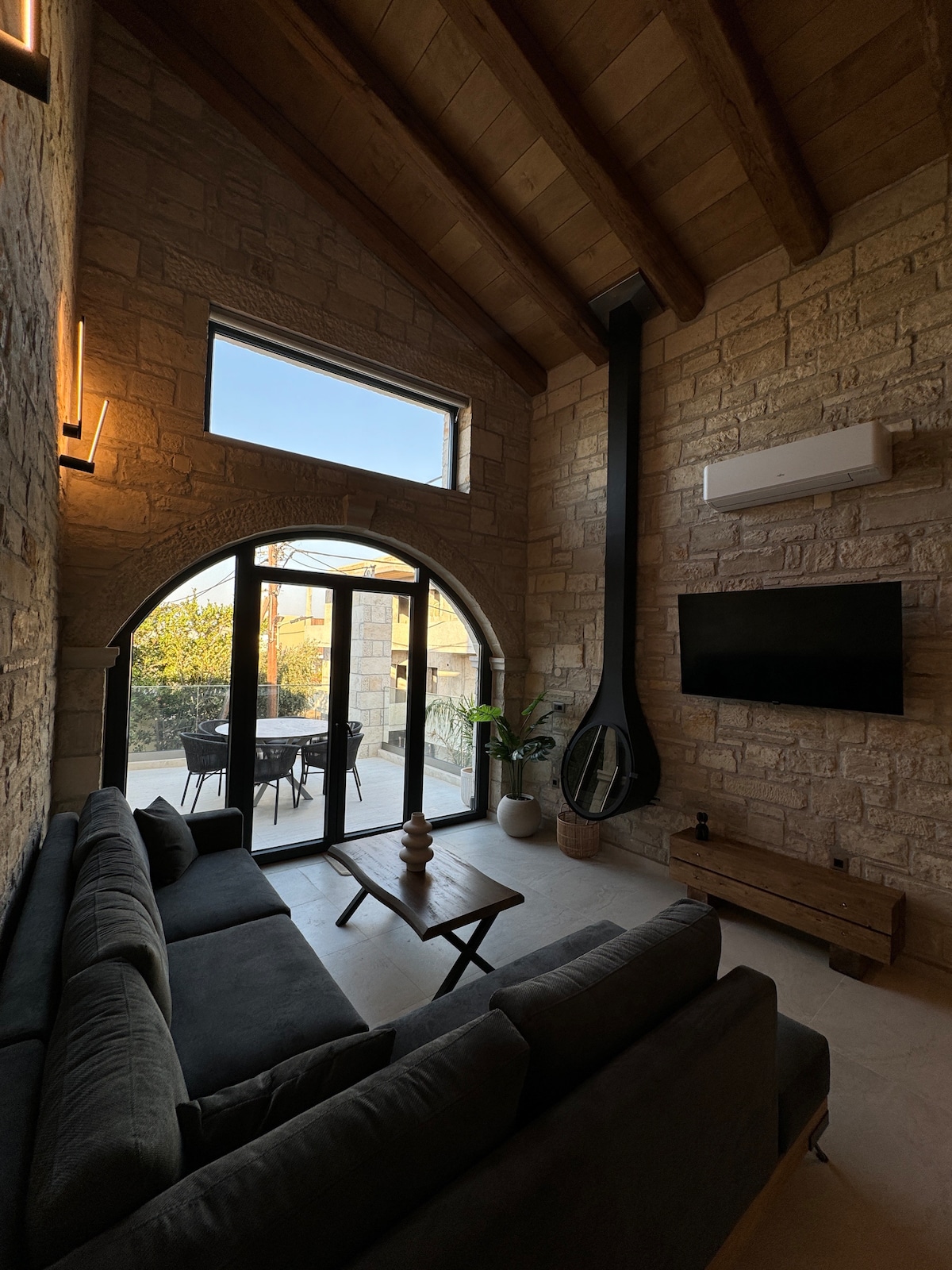 A spacious living area features high wooden ceilings and a large, arched window. A dark sofa is arranged in front of a modern television, while a small coffee table sits nearby. Natural light fills the room, enhancing the stone walls and indoor plants.