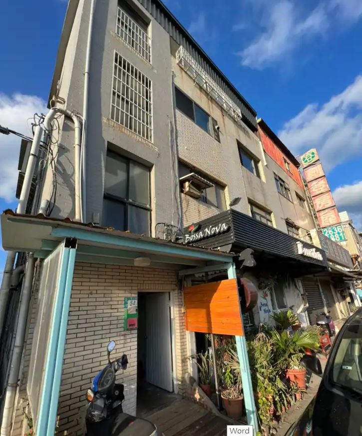 A multi-story building features a blend of textured gray and bright awning finishes. Large windows with metal grilles line the exterior, while greenery is visible near the entrance. Nearby, a parked motorcycle adds to the urban setting under a partly cloudy sky.