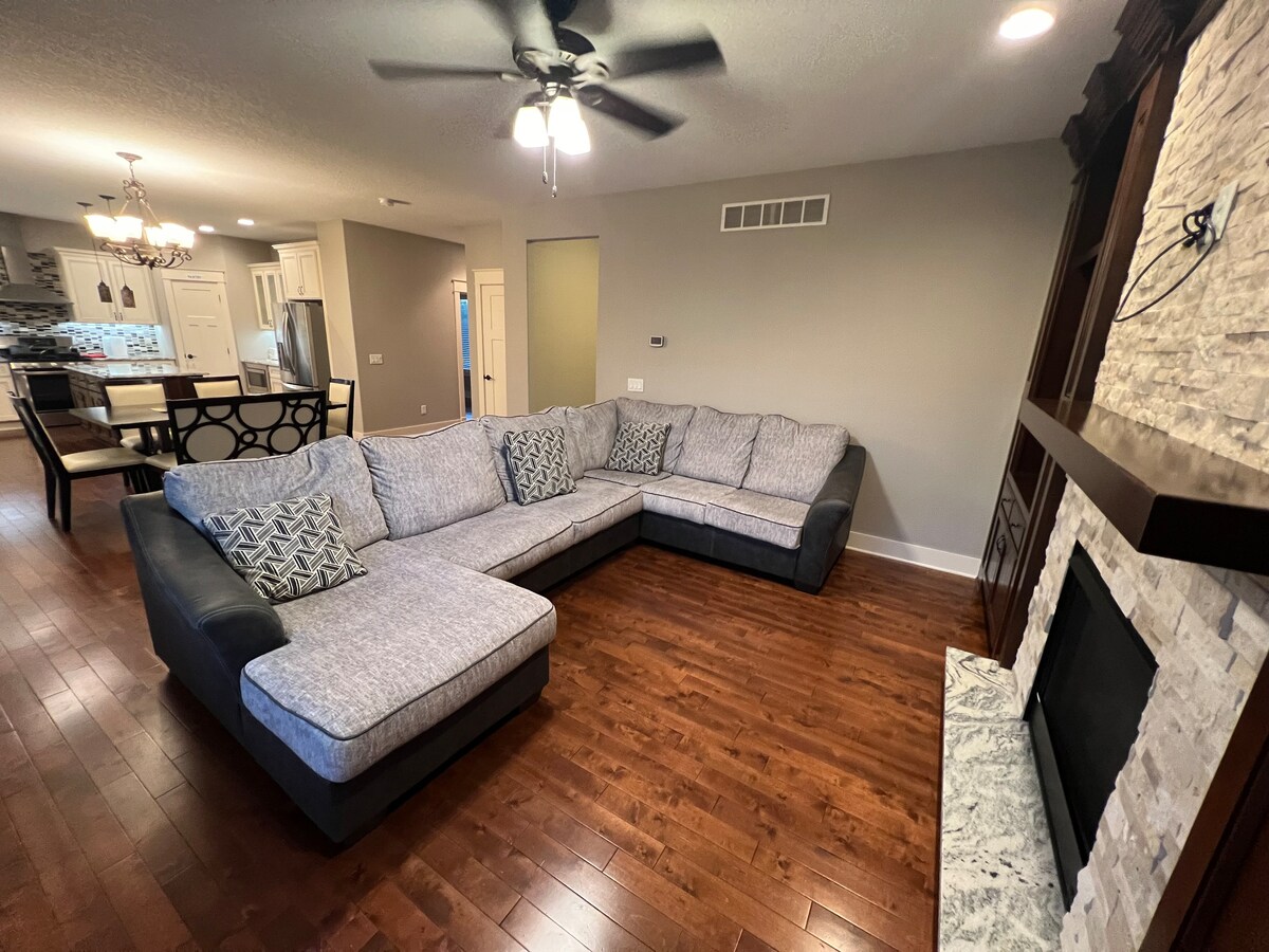 A spacious living area features a large sectional sofa with decorative pillows. A ceiling fan hangs above, and light wood flooring complements the room's neutral-colored walls. A cozy fireplace is located on one side, with a dining area visible in the background.