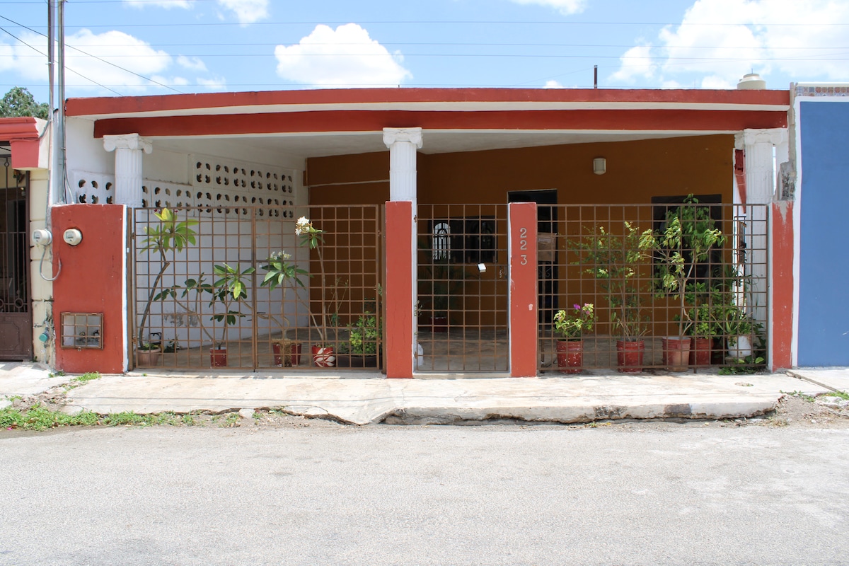 The exterior of the property is presented, featuring a red and white façade with a gated entrance. Planters with greenery line the front, creating a welcoming appearance. The structure is single-story, with overhead clouds visible in the blue sky above.