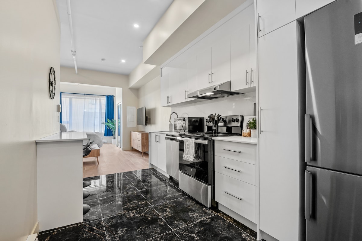A modern kitchen is displayed, featuring sleek white cabinetry and stainless steel appliances, including a refrigerator and oven. A glossy black marble floor reflects light, while a dining area with minimalist chairs is visible in the background. Natural light enters through a sliding glass door.
