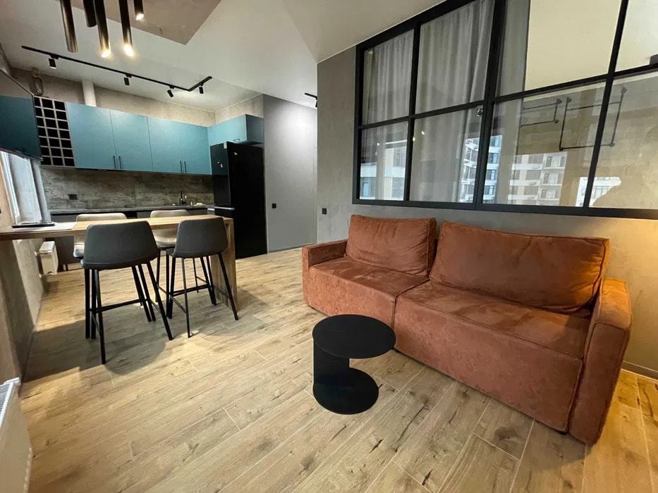 The living area features a spacious, orange sofa paired with a small black coffee table. A dining set for four is positioned nearby. The kitchen area showcases modern cabinetry in muted blue tones, accompanied by large windows that allow natural light to fill the space.